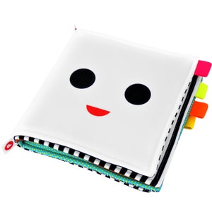 May include: A white fabric book with a black and white patterned border. The front cover features a simple black and white face with a red smile. The book has colorful fabric tabs on the side.