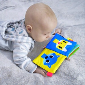 May include: A baby in a gray and white striped hooded onesie is looking at a colorful soft book with animal faces. The book has yellow, blue, green, and orange pages.
