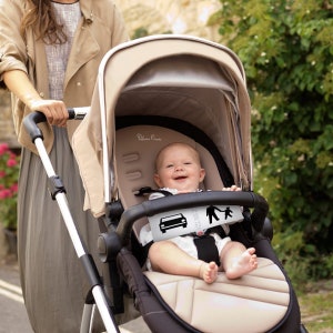 May include: A beige stroller with a black frame and a baby sitting inside. The stroller has a black handle and a black canopy. The baby is wearing a white shirt and has a happy expression on their face.
