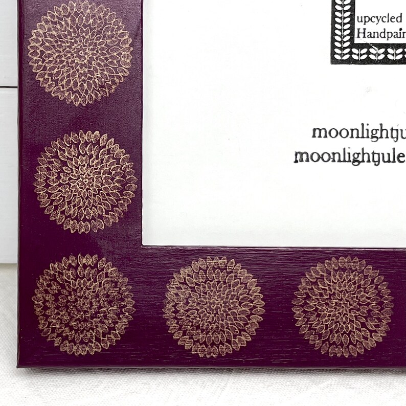 May include: A purple picture frame with a gold floral design. The frame has a white mat and the text "upcycled Handpainted moonlightjule moonlightjule" is printed on the mat.