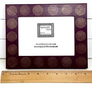 May include: A dark purple picture frame with a gold floral design. The frame is 8 inches wide and has a white mat. The frame is made by Moonlight Jules, a company that makes hand-painted frames.
