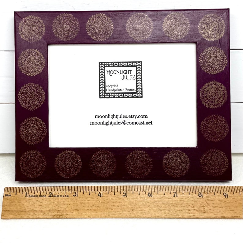 May include: A dark purple picture frame with a gold floral design. The frame is 8 inches wide and has a white mat. The frame is made by Moonlight Jules, a company that makes hand-painted frames.