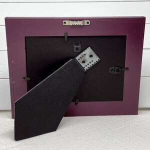 May include: A purple picture frame with a black mat and a black easel stand on the back. The frame has a metal hanging hook on the back.