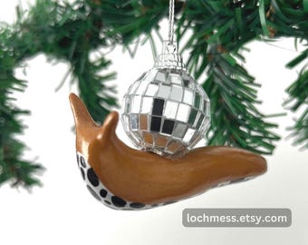 Puede incluir: Un adorno de caracol marrón y negro con manchas que sostiene un adorno de Navidad de bola de discoteca plateada. El adorno está colgado de un árbol de Navidad artificial verde.