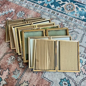 Puede incluir: Una colección de marcos de fotos rectangulares con bisagras y bordes metálicos dorados finos. Los marcos están dispuestos en una formación escalonada y superpuesta, con frentes de cristal y varios insertos que incluyen papel blanco liso y cartón texturizado.