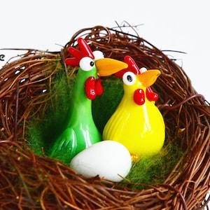 May include: Two glass chickens, one green and one yellow, are sitting in a brown woven nest with a white egg. The chickens have red combs and beaks, and large black eyes.