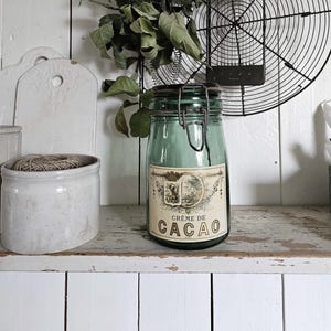 Pode incluir: Uma cena de cozinha em estilo vintage com um frasco de vidro verde rotulado "Crème de Cacao". O frasco, selado com um fecho de metal, está numa prateleira de madeira. Um molho de folhas verdes está pendurado por cima, juntamente com uma grelha de arame e outros acessórios de cozinha.