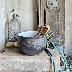 May include: A vintage still life composition. A black and white enamel pot with wooden spoons, a retro seltzer bottle, and wooden eggs are arranged on a weathered wooden surface. A distressed wooden door with a decorative element is in the background.