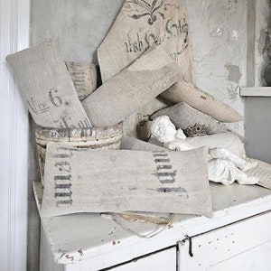 Peut inclure: Une collection d'oreillers en lin de style vintage avec des lettres noires imprimées et un sac en toile de jute avec des motifs floraux noirs et des lettres. Les oreillers et le sac sont disposés sur une surface peinte en blanc avec un seau en bois et deux figurines de chérubins.