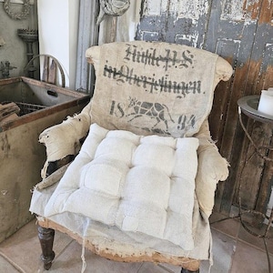 Könnte beinhalten: Ein Vintage-Sessel mit einem abgenutzten, weißen Stoffkissen und einem Jutesack mit dem aufgedruckten Text "Hütte 18 94". Der Sessel steht in einer rustikalen Umgebung mit einer Holztür und einem Metallstern.