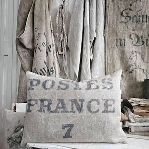 May include: A rectangular linen pillow with the words "POSTES FRANCE 7" printed in dark gray. The pillow is on a white wooden surface, with other linen textiles hanging in the background. The overall aesthetic is rustic and vintage.