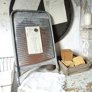 May include: A vintage metal washboard with a rusty patina, leaning against a black circular metal object.  The washboard has a series of raised ridges and is attached to a wooden frame.  There are three pieces of paper with handwritten text attached to the metal object with magnets.  A wooden box with a rustic finish contains several bars of soap.