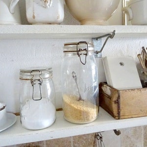 Op de afbeelding: Witte keukenkasten met verschillende witte keramische items, waaronder kommen, potten en een kan. Twee glazen potten met metalen deksels zijn gevuld met witte en gele korrelige stoffen. Een houten doos met een metalen handvat bevindt zich op de onderste plank.