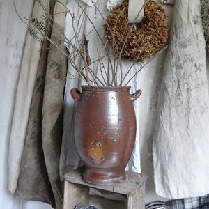 Può includere: Un grande vaso di terracotta marrone si trova su una cassa di legno davanti a un muro decorato con tessuto bianco e ghirlande di fiori secchi.