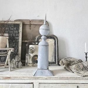 Op de afbeelding: Een verweerd grijs metalen sierstuk met een puntige top en een ronde basis. Het sierstuk staat op een witte houten tafel met andere decoratieve items.