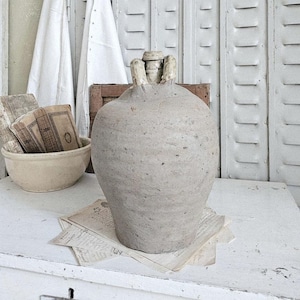 May include: A large, textured, gray ceramic jug with a handle sits on a white surface. The jug is surrounded by vintage papers and a small bowl. White fabric hangs in the background, creating a rustic, antique aesthetic.