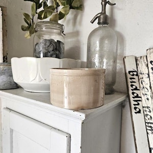 Può includere: Un vaso cilindrico in ceramica beige è appoggiato su un mobile bianco. Dietro si trovano una ciotola bianca smerlata contenente un barattolo di vetro pieno di gettoni metallici, un sifone per soda in vetro trasparente e insegne in legno con il testo Buddha.