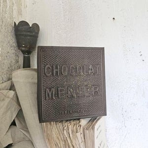 Könnte beinhalten: Ein quadratisches, dunkelbraunes Metallschild mit einer texturierten, gitterartigen Oberfläche. Der geprägte Text lautet "CHOCOLAT MENIER" in großen Großbuchstaben, wobei das kleinere Wort "TABLETTES" darunter zu sehen ist. Das Schild wirkt verwittert.