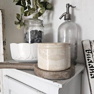 Può includere: Una natura morta con un sifone per soda vintage, un piccolo barattolo di ceramica e un barattolo di vetro pieno di piccoli oggetti rotondi. Sono visibili anche una ciotola decorativa bianca e un'insegna di legno con la parola "Buddha". Foglie verdi sono appese sopra i barattoli.