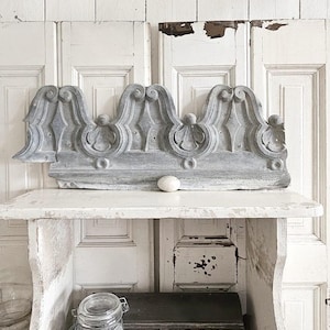 May include: A distressed, grey-painted architectural salvage piece with decorative scrollwork is displayed on a white shelf. A small, white, oval stone sits on the shelf. A glass jar and a dark metal box are also visible.