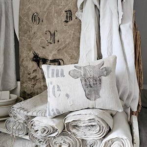 May include: A stack of rolled white fabric with a pillow featuring a cow design and the text "No. 1. G. D. 2f." on a burlap background.