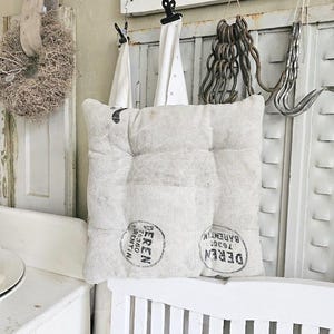 May include: A square, off-white cushion with a tufted design and a vintage aesthetic. The cushion has two circular black stamps with text. It rests on a white slatted bench, with other vintage decor in the background.