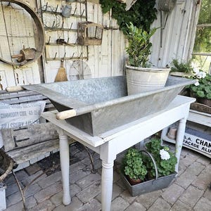 Può includere: Una vasca in metallo zincato con manici in legno è appoggiata su un tavolo bianco. Una pianta in vaso è all'interno della vasca. Il tavolo è su un patio in mattoni, con altri oggetti vintage sullo sfondo, creando un'estetica rustica.