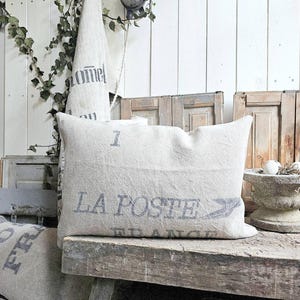 May include: A rectangular, off-white pillow with the text "LA POSTE FRANCE" in faded blue lettering. The pillow is made of a textured fabric and rests on a weathered wooden surface. A matching towel with text hangs in the background.