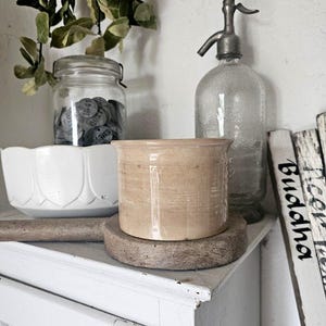 Può includere: Una natura morta con un sifone per soda in vetro, un piccolo vaso in ceramica beige (circa 10 cm di diametro) su un sottobicchiere in legno e una ciotola bianca. È presente anche un barattolo di vetro pieno di oggetti grigi e vegetazione.