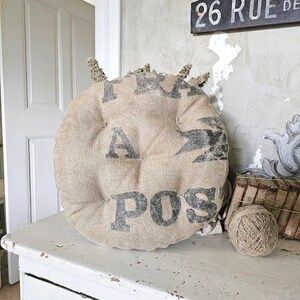 May include: Round, beige burlap cushion with black stenciled text. The cushion is placed on a distressed white wooden surface. A ball of twine and a small wooden basket are also visible. The cushion has a rustic, vintage aesthetic.
