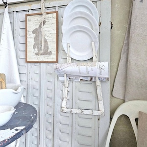 May include: A rustic display featuring white plates, a framed bunny print, and a vintage-style newspaper roll. The plates are stacked on a distressed white rack, adding a touch of farmhouse charm to the scene.