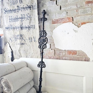Op de afbeelding: Een decoratieve zwarte metalen staaf met sierlijke bloemen- en krulwerkdetails staat tegen een verweerde bakstenen muur. Een vintage linnen banner met vervaagde tekst hangt in de buurt en rollen stof zijn eronder gestapeld.