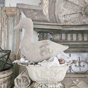 Può includere: Un'anatra decorativa in tessuto beige chiaro, appoggiata in una fioriera in metallo bianco anticato. La fioriera presenta un design ornato e dettagliato, ed è posizionata su una superficie di legno invecchiato. L'estetica generale è vintage e rustica.