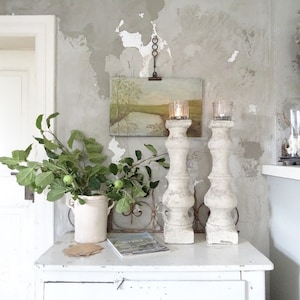 May include: Two white decorative candle holders with a rustic finish, a small painting of a landscape, and a white ceramic vase with green leaves and small green apples on a white distressed dresser with two drawers.