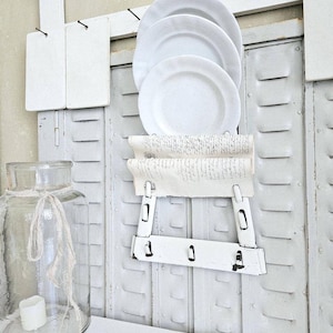 Puede incluir: Un estante blanco rústico para platos con cuatro platos blancos. El estante está montado sobre una superficie metálica blanca desgastada. Un tarro de cristal transparente con una vela y una cinta está cerca.