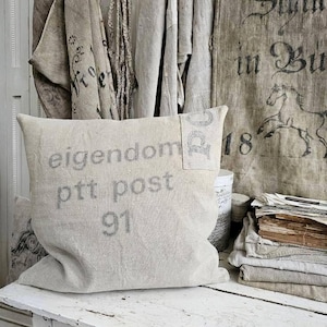 May include: A beige linen pillow with the words "eigentdom ptt post 91" printed in gray. The pillow is on a white, distressed wooden table. Other linen textiles and a vintage-style wall hanging are in the background.