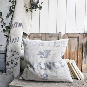 May include: A linen pillow with faded blue text reading "Postes France 2" sits on a weathered wooden surface. A matching linen towel with text hangs in the background, alongside a vintage-style light fixture and greenery.