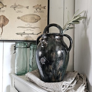 May include: A dark, glazed ceramic vase with two handles and a spout, resting on a linen cloth. The vase is placed on a white shelf, with two glass jars and a vintage fish chart in the background. A sprig of greenery is placed in the vase.