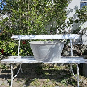 May include: A weathered, white bench with a galvanized metal tub. The bench has a distressed finish, and the tub has two handles. The scene is outdoors, with greenery in the background.