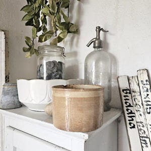 Può includere: Un vaso cilindrico in ceramica color marrone chiaro si trova davanti a una ciotola bianca smerlata, un barattolo di vetro pieno di monete metalliche e un sifone per soda in vetro. A destra, tre listelli di legno bianco riportano testo nero, tra cui Buddha.
