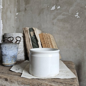 May include: A white ceramic jar with a rounded body and a slightly flared rim sits on a piece of paper atop a weathered wooden surface. Behind it are old books and a small metal bucket.