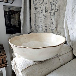 May include: A cream-coloured ceramic bowl with a scalloped rim and a thin brown edge detail. The bowl has a smooth, rounded interior and a slightly textured exterior surface, showcasing a simple, functional design for serving or display purposes.