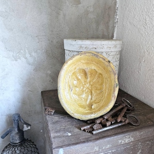 Puede incluir: Naturaleza muerta vintage con un cuenco de cerámica amarilla con diseño floral, un recipiente cilíndrico blanco con escritura y una colección de llaves antiguas. A la izquierda se ve una botella de vidrio envuelta en alambre.