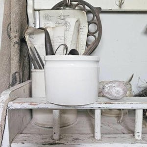 Puede incluir: Un tarro de cerámica blanca sobre una estantería de madera pintada de blanco envejecido. La estantería exhibe varios objetos vintage, incluyendo papeles antiguos, utensilios y una pieza metálica decorativa. Una tela de arpillera cuelga a la izquierda.