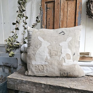 Op de afbeelding: Een vierkant kussen van grof, lichtbruin textiel met twee witte konijnensilhouetten als applicatie op de voorzijde. Het kussen vertoont zichtbare stiksels, rafelige randen en donkere, vervaagde tekstmarkeringen op het gestructureerde oppervlak.