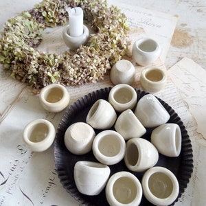 May include: A collection of small white ceramic pots arranged in a black metal tray with a scalloped edge. The pots are arranged in a circular pattern with a few pots overlapping. The pots are all the same size and shape, and they have a smooth, white finish.