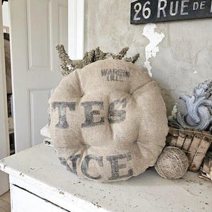May include: A round, beige cushion with the words "TES RICE" printed in dark gray. The cushion is placed on a distressed white wooden surface, with a ball of twine and decorative items nearby. The cushion has a vintage, farmhouse aesthetic.