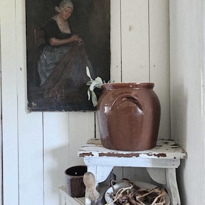 May include: A brown ceramic crock with side handles sits on a small white distressed wooden stool. Below, a white bowl holds several brown onion bulbs with dried roots. A framed oil painting of a person in a white cap hangs on the wall.