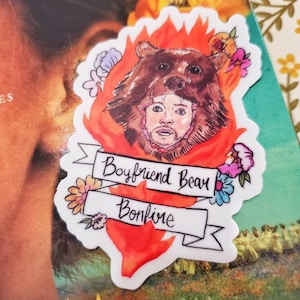May include: A sticker with a watercolor illustration of a man wearing a bear head. The bear head is on fire and there are flowers around the man's head. The sticker says "Boyfriend Bear Bonfire".