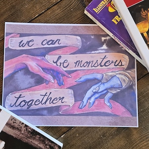 May include: Print featuring two hands reaching towards each other, with the text "we can be monsters together" on pink banners. The artwork has a gothic theme with a dark, moody color palette. Books are in the background.